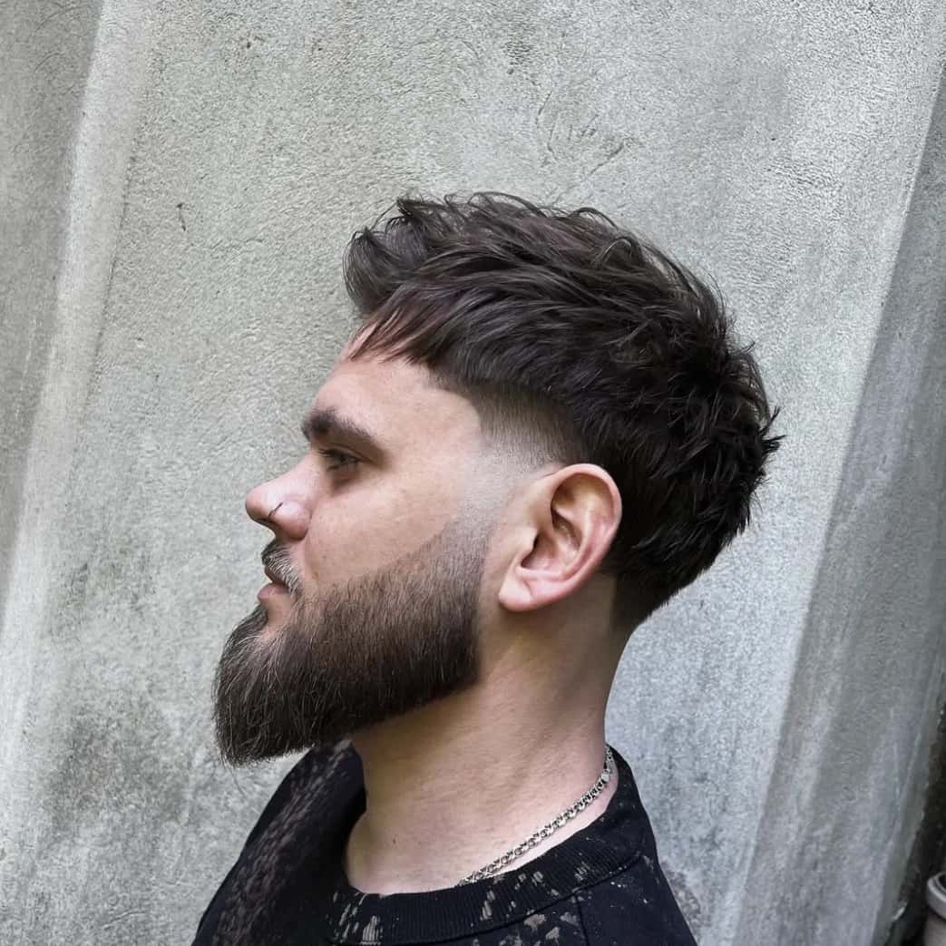 A textured crop with a fade and a full beard, in profile against a concrete wall